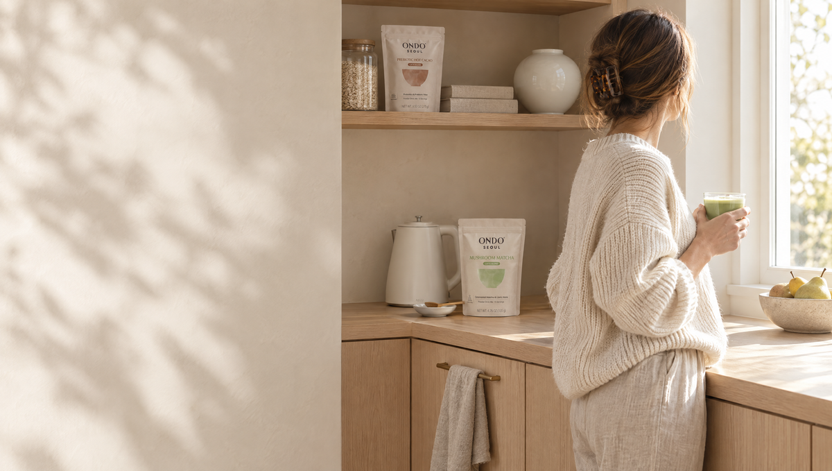 A woman holding an iced matcha latte in a bright kitchen, with ONDO° pouches on the counter and shelf.