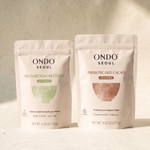 Day & Night Duo: two pouches, Focus Mushroom Matcha Latte Blend and Prebiotic Hot Cacao Latte Blend, on ivory hanji paper.