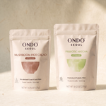 The Long Day Duo: two pouches, Prebiotic Matcha Latte Blend and Focus Mushroom Hot Cacao Latte Blend, on ivory hanji paper.